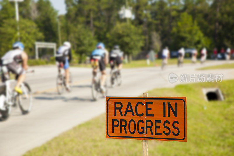 一大群骑自行车的人在比赛。“Race in Progress”标志着前景。